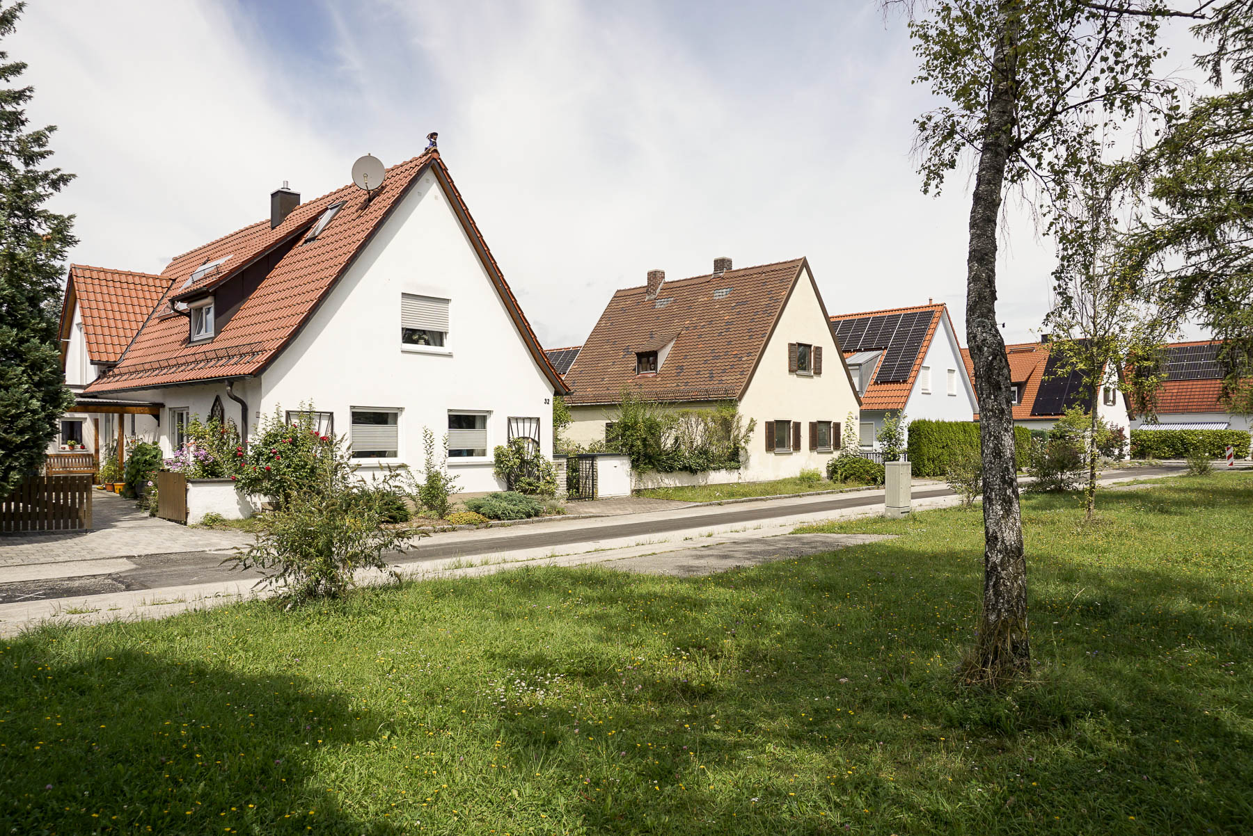 Immobilienmakler Gilching: Blick auf ein weißes Einfamilienhaus mit roten Ziegeldach und Solaranlage in einer ruhigen, grünen Wohnstraße.