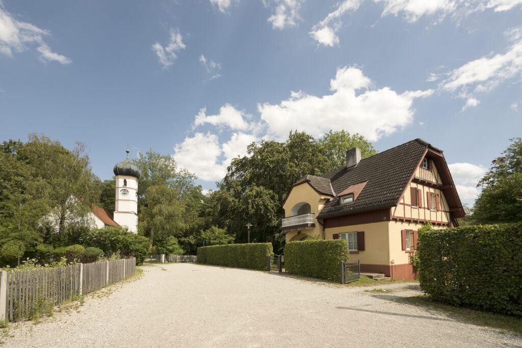 Immobilienmakler Krailing: Idyllischer Weg in Krailling mit einem traditionellen Haus mit Fachwerk und Balkon auf der rechten Seite sowie einer Kirche mit Zwiebelturm im Hintergrund.