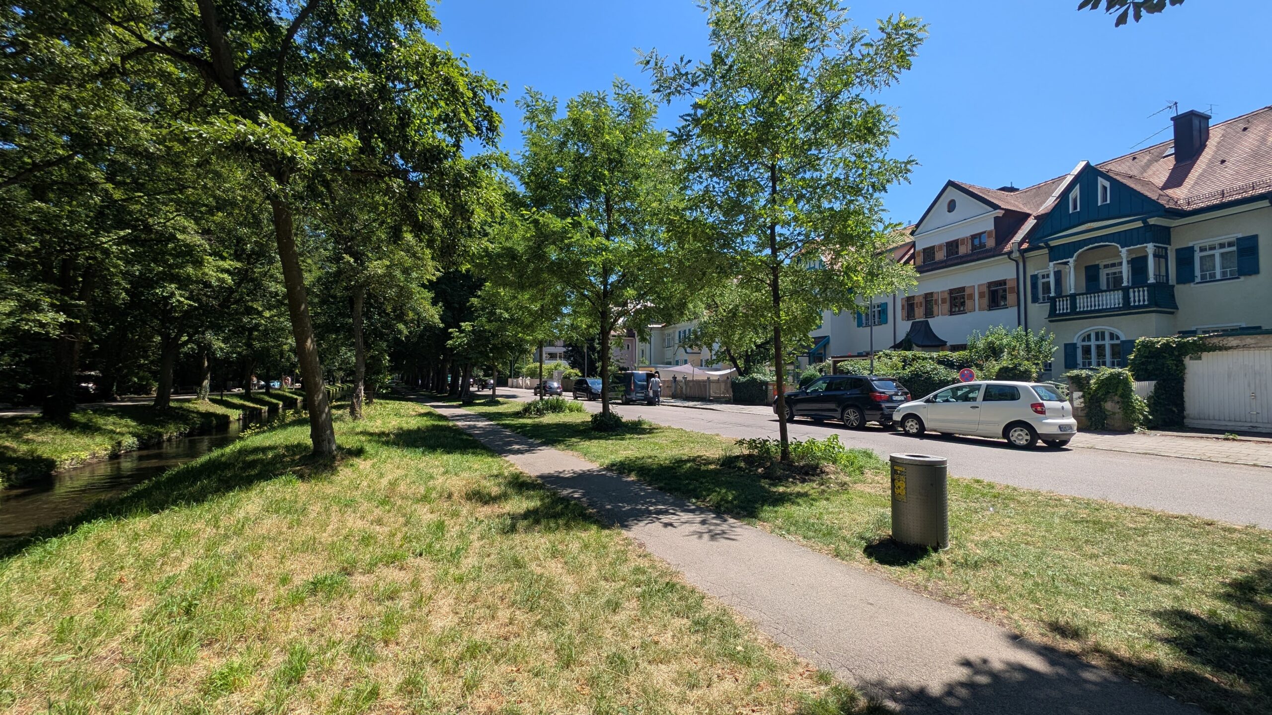 Sonnige Straßenszene an der Marsopstraße in Pasing mit einem schmalen Bachlauf und viel Grün auf der linken Seite, einem Fußweg sowie repräsentativen Häusern, parkenden Autos und Bäumen auf der rechten Seite unter blauem Himmel.