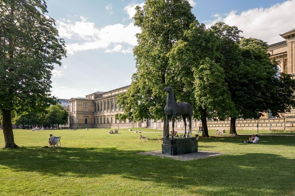 Immobilienmakler Maxvorstadt: Blick auf die Alte Pinakothek in München Maxvorstadt mit einer großen Grünfläche, Bäumen, erholenden Menschen und einer bronzenen Pferdeskulptur im Vordergrund