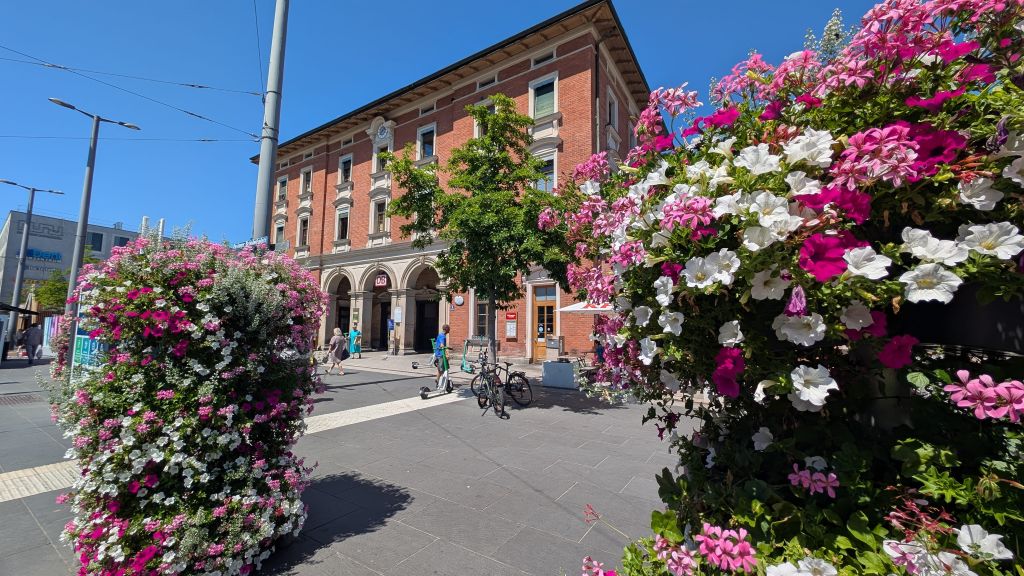 Immobilienmakler Pasing: Der Bahnhofsplatz in Krailling mit einem historischen roten Backsteingebäude im Hintergrund. Im Vordergrund rahmen große, üppige Blumenampeln in Rosa und Weiß den Weg ein. Passanten gehen über den Platz, und Fahrräder sind abgestellt.
