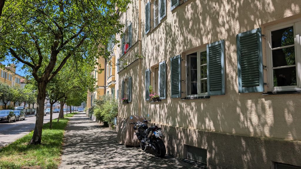 Immobilienmakler Pasing: Blick auf eine helle Wohnhausfassade mit blauen Fensterläden in Krailling an einem sonnigen Tag. Auf dem Gehweg, der von Bäumen gesäumt ist und Schatten wirft, steht ein Motorrad unter einer Abdeckung. Einige Autos parken auf der Straße.