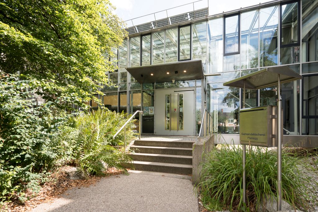 Immobilienmakler Planegg: Eingangsbereich der modernen Gemeindebücherei in Planegg mit einer Glasfassade, einer Treppe und einem Briefkasten im Vordergrund, umgeben von Bäumen und Sträuchern.