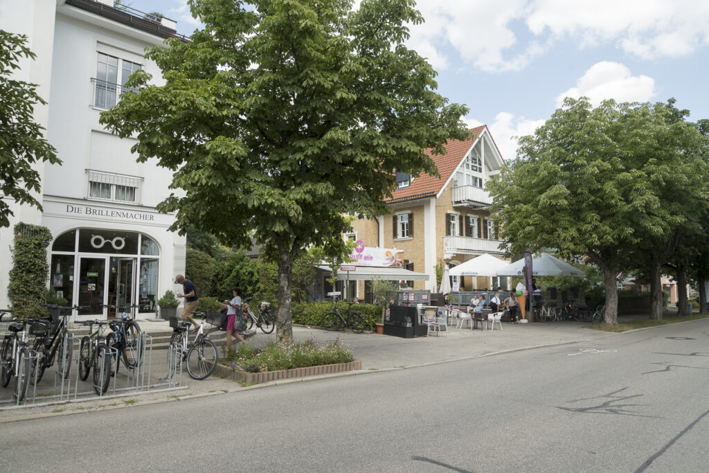 Immobilienmakler Planegg: Ein belebter Straßenzug in Planegg mit einem Optikergeschäft, abgestellten Fahrrädern und einem Café mit Außengastronomie unter Sonnenschirmen, gesäumt von großen grünen Bäumen.