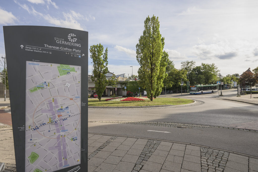 Immobilienmakler Germering: Eine große Infotafel mit Stadtplan im Vordergrund zeigt Wege und Orte in Germering. Im Hintergrund ist ein begrünter Kreisverkehr mit Blumenbeeten, schlanken Bäumen und einer vorbeifahrenden Buslinie zu sehen.