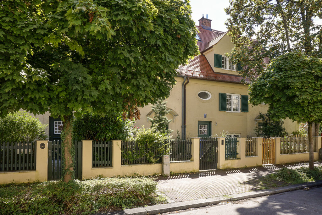 Immobilienmakler Laim: Eine charmante, von Bäumen gerahmte Wohnimmobilie mit einer warmen Fassade, traditionellen grünen Fensterläden und einem gepflegten Holzzaun in einer ruhigen Nachbarschaft von Laim.