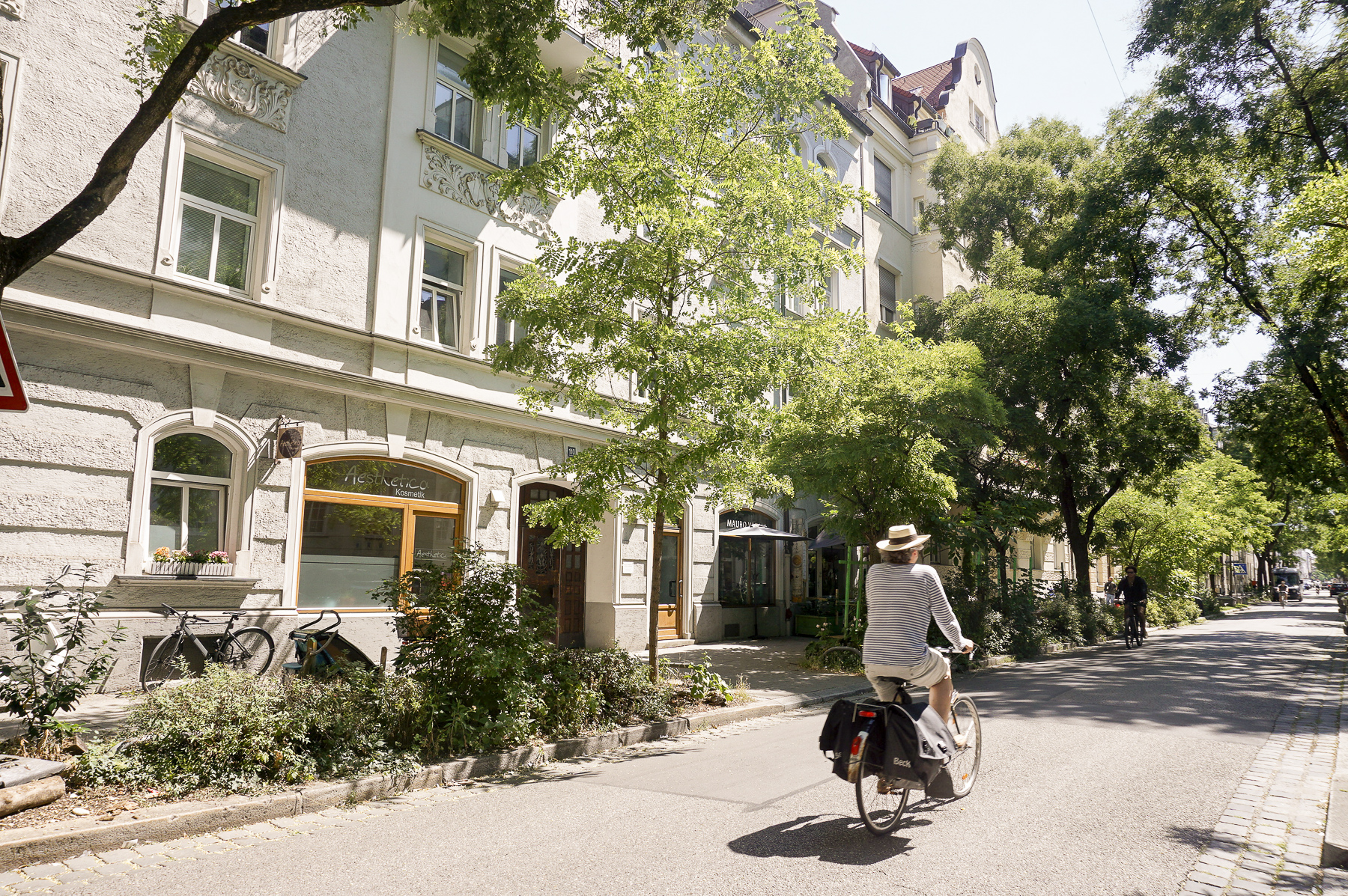 Immobilienmakler Neuhausen: Eine sonnige Straßenszene, in der eine Person gemütlich mit dem Fahrrad entlang historischer Wohngebäude und üppigem Grün fährt.